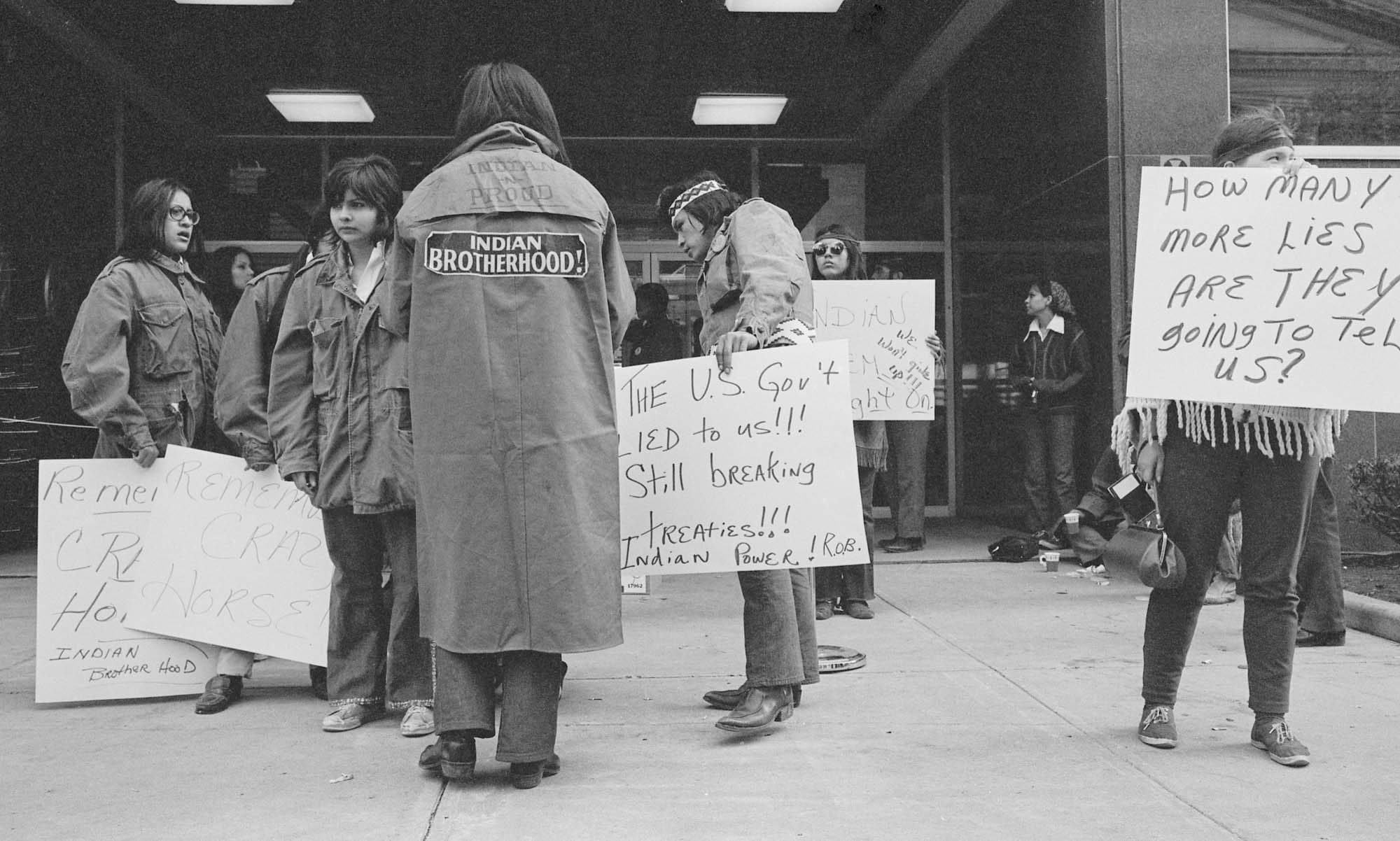 Provided by Minnesota Historical Society Press. Photos by Dick Bancroft ‚ A group of AIM women protest at the front door of the US Courthouse in Minneapolis. This is a black-and-white photo of people holding signs outside the courthouse. One with back to camera wears a coat with sign on back saying‚”Indian Brotherhood.”